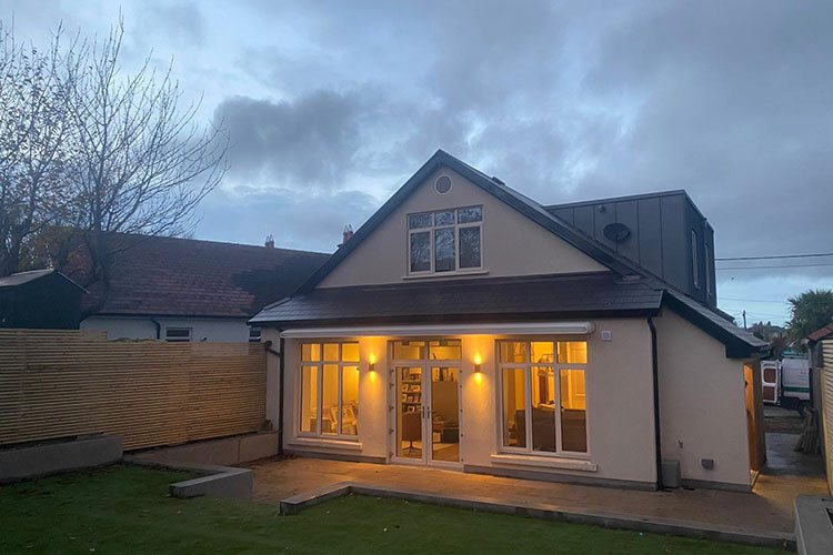 A house at dusk with warm light in the windows of a rear extension, seen across the back lawn